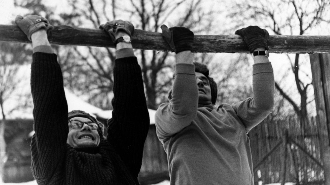 Кадр з фільму: двоє чоловіків у шапках і свертах підтягуються на колоді вгору