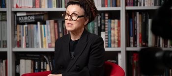 Токарчук сидить у кріслі, на тлі неї бібліотека з книжками