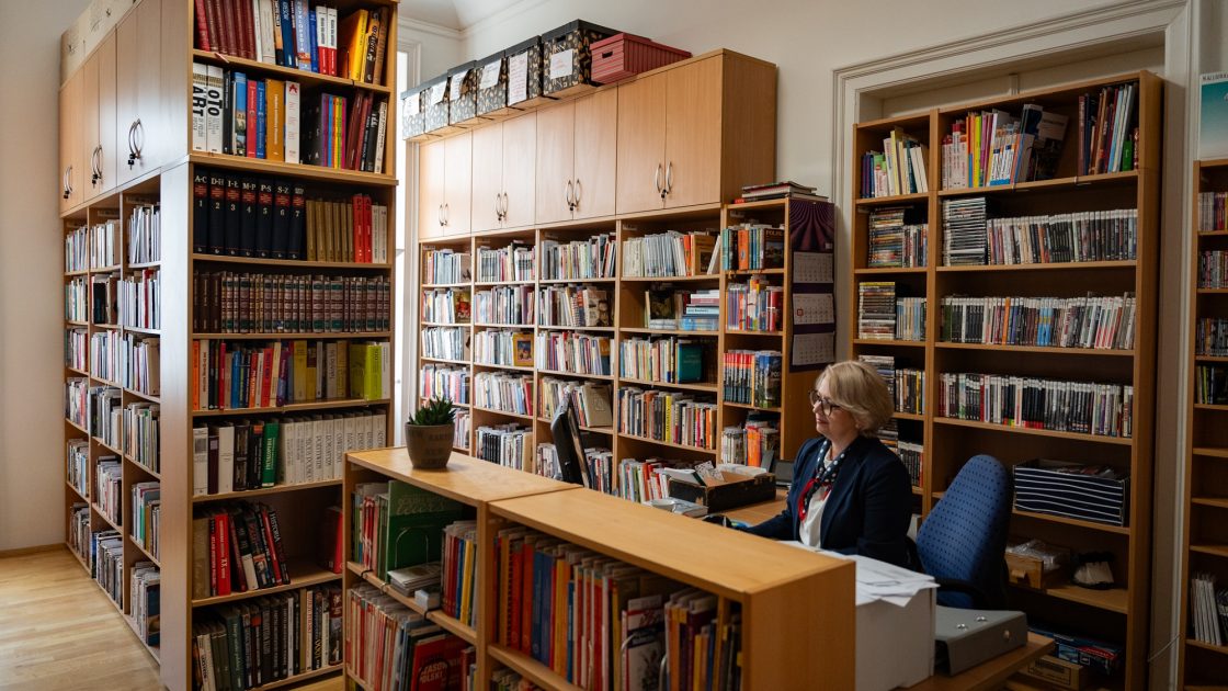 Na fotografii je knihovna Polského institutu - regály s knihami a v popředí stůl paní knihovnice, která sedí za monitorem počítače.
