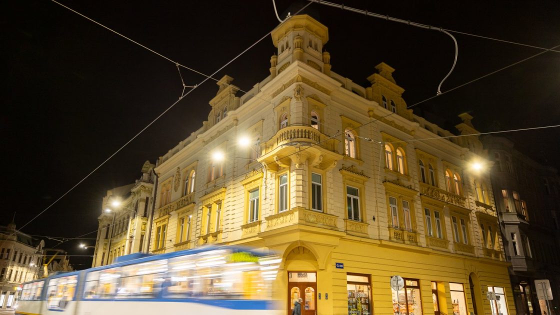 Rohový měšťanský dům v temné ulici, před ním jede tramvaj. Dům je žlutě nasvícený a vypadá reprezentativně.