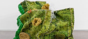 Fotografie exponátu z výstavy Deep House. Na bílém pozadí se nachází objekt ve tvaru křesla s područkami, je vytvořený ze starých koberců zelené barvy.