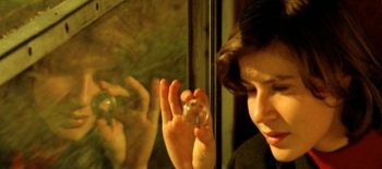 Na fotografii - záběru z filmu Dvojí život Veroniky je mladá žena, která se dívá z okna vlaku. Fotografie zachycuje také její odraz v okně. V ruce drží skleněnou kuličku, do které hledí skrz sluneční paprsky.