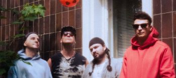 Na fotografii stojí čtyři muži ve sportovním oblečení, dva z nich mají sluzeční brýle. Za nimi je okno domu. A nad nimi se vznáší míč.