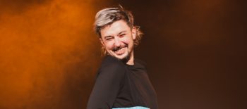 Fotografie z divadelního představení Pan Tadeáš představuje usměvavého mladého muže s vousy a účesem s částečně odbarvenými vlasy. Muž je oblečený do tmavého oděvu, přes který má modrý dámský korzet.