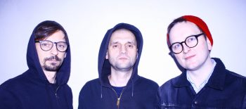 Fotografie členů skupiny Trupa Trupa, tři muži středního věku v tmavém oblečení, na světlém pozadí.