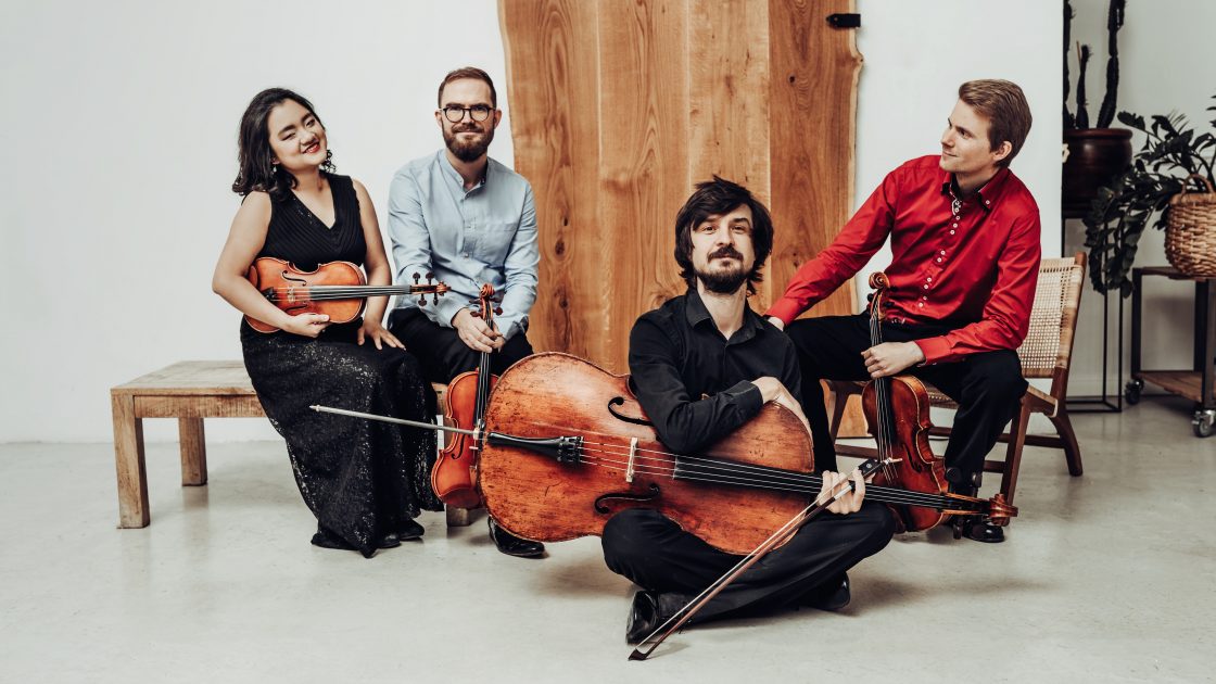 Na obrázku jsou čtyři mladí hudebníci - členové kvarteta, mají u sebe své hudební nástroje: housle, violu či violoncello.