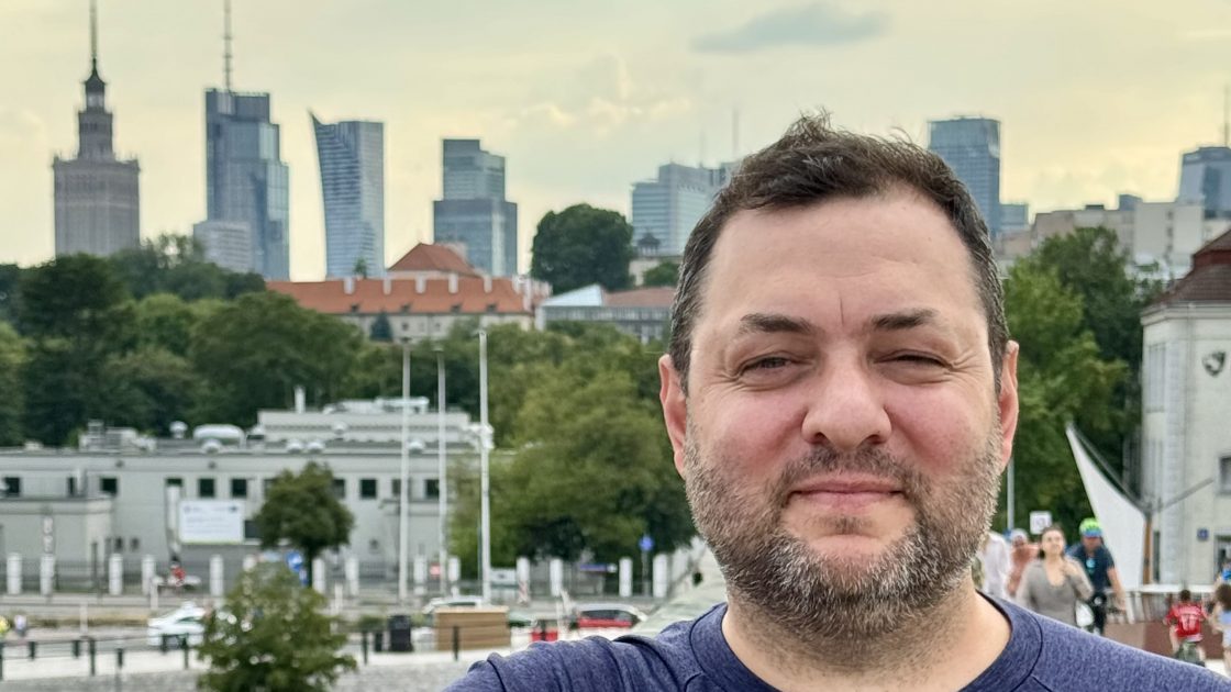 Na fotografii je Pavel Trojan - usměvavý muž středního věku, který se opírá o zábradlí a v pozadí jsou vidět varšavské mrakodrapy.