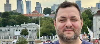 Na fotografii je Pavel Trojan - usměvavý muž středního věku, který se opírá o zábradlí a v pozadí jsou vidět varšavské mrakodrapy.
