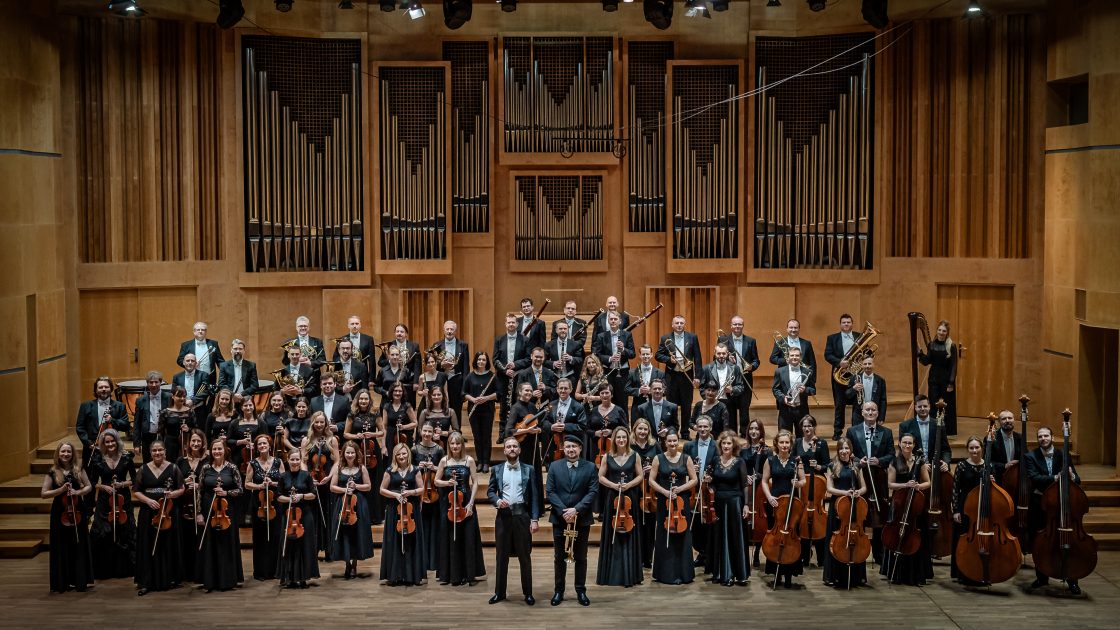 Na fotografii je symfonický orchestr z města Opolí. Skupinová fotografie velkého orchestru je v interiéru koncertní síně, v pozadí jeviště jsou vidět píšťaly varhan.