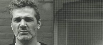 Na fotografii je hudebník Robert Piotrowicz, muž středního věku v tmavém oblečení. Snímek je pořízen s pozadím starého okna s mříží nebo sítí.