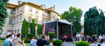 Na fotografii je barokní budova Fürstenberského paláce a v jeho zahradě je umístěno pódium, před kterým sedí na trávě lidé