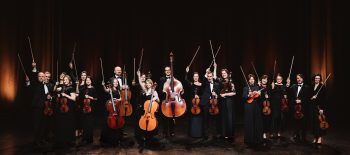 Na fotografii je smyčcový orchestr, hudebníci drží v rukou své nástroje a zdvihají smyčce do výšky.