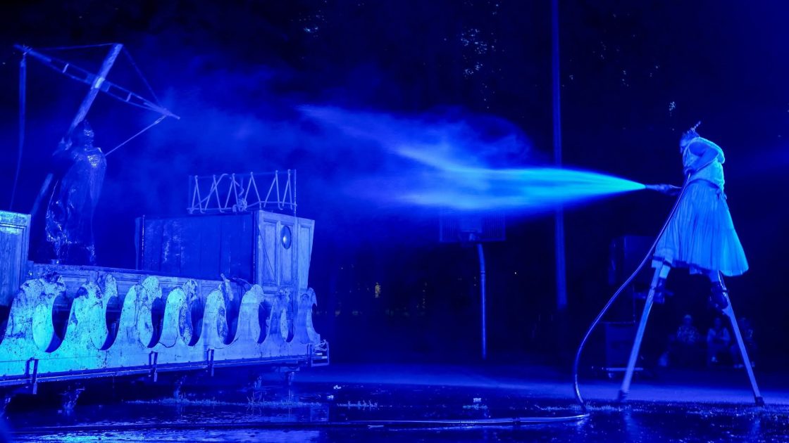 Na obrázku je osoba na chůdách. Scenérie z divadelního představení Bouře je ve tmě, modře nasvícená.