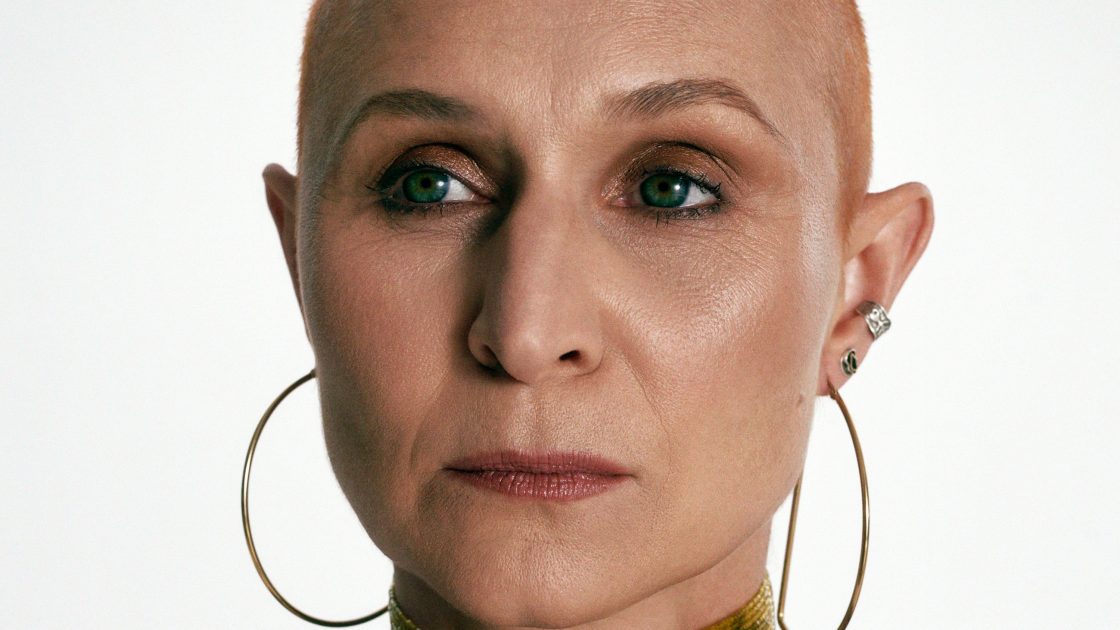 Žena středního věku s výrazným líčením a etnickými šperky, má celké kruhové náušnice a několik náhrdelníků. Má vlasy obarvené do téměř oranžové barvy a po stranách vyholené, účes podobný punku.