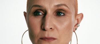 Žena středního věku s výrazným líčením a etnickými šperky, má celké kruhové náušnice a několik náhrdelníků. Má vlasy obarvené do téměř oranžové barvy a po stranách vyholené, účes podobný punku.