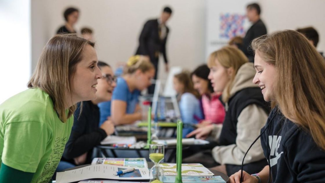 Lidé (převážně ženy) sedící u stolků ve dvojicích a hovořících navzájem. Na stolcích jsou nápisy s různými jazyky.