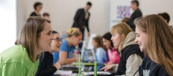 Lidé (převážně ženy) sedící u stolků ve dvojicích a hovořících navzájem. Na stolcích jsou nápisy s různými jazyky.