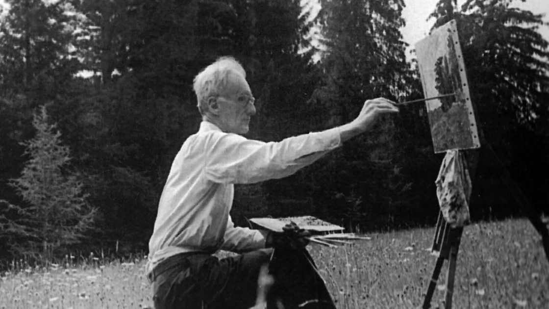 Černobílá fotografie staršího muže, který sedí venku na louce u lesa na přenosné stoličce u malířského stojanu a maluje obraz - krajinu.