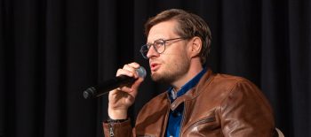 Muž mladšího středního věku v brýlích, v kožené bundě, sedí na židli a v ruce má mikrofon
